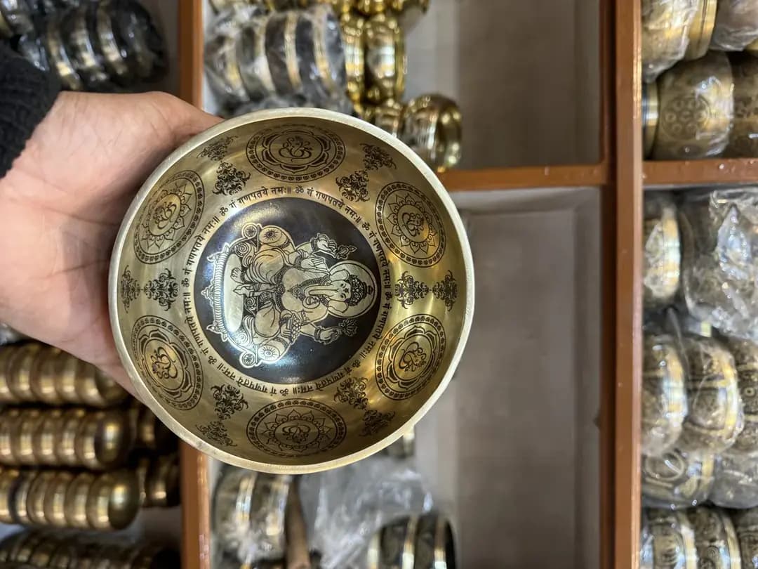 An artisan holding a hand-etched bronze singing bowl, shelves of finished pieces behind
