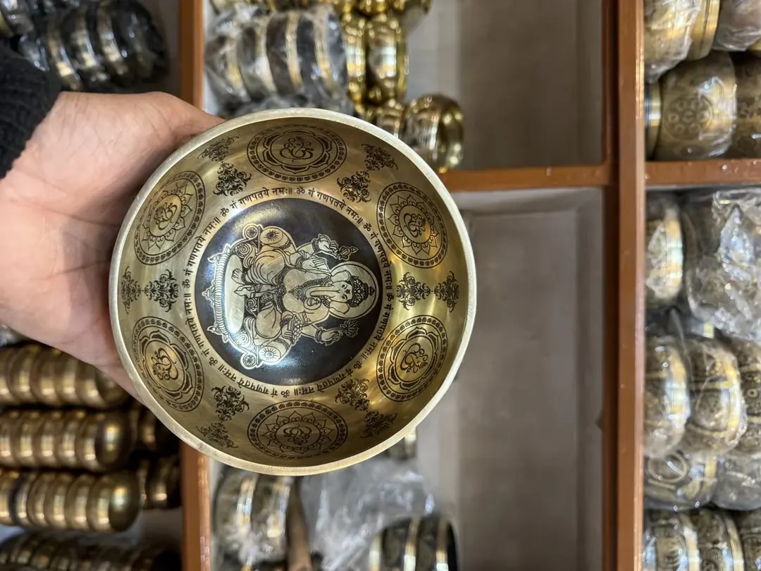 An artisan presenting a hand-etched bronze singing bowl in a Patan workshop, shelves of finished bowls behind